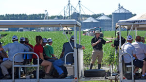 An NC State Extension specialist talking into a microphone in a soybean field while giving a tour to participants sitting in a tram.
