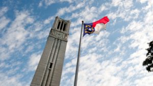 The North Carolina flag atop a flag pole beside the NC State University belltower, with a blue sky and white clouds above.